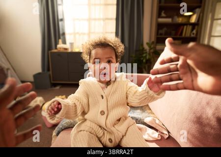 Enfant nourrisson avec des cheveux bouclés assis sur le canapé atteignant les mains tendues de l'homme noir dans le cadre de la maison, illustrant le lien et les soins pendant le congé de paternité Banque D'Images