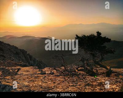 Coucher de soleil sur le paysage de montagne de la Crète, Grèce, avec le ciel dramatique et la silhouette d'un arbre tordu sur un terrain rocheux. Banque D'Images