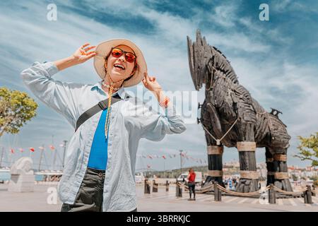 6 mai 2025, Canakkale, Turquie : heureuse touriste ajustant son chapeau tout en posant près de la réplique du cheval de Troie, par une journée ensoleillée Banque D'Images
