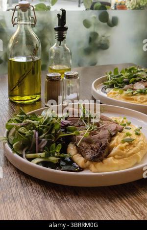 Une assiette gourmande avec des tranches de rôti de bœuf sur une purée de pommes de terre crémeuse, accompagnée de légumes verts frais mélangés et de bouteilles d'huile d'olive sur un ta en bois rustique Banque D'Images