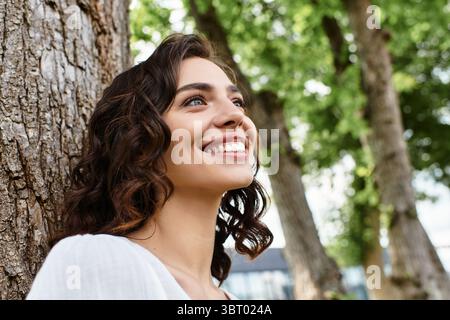 Une jeune femme aux cheveux bruns ondulés sourit joyeusement tout en profitant d'une journée ensoleillée dans le parc. Banque D'Images