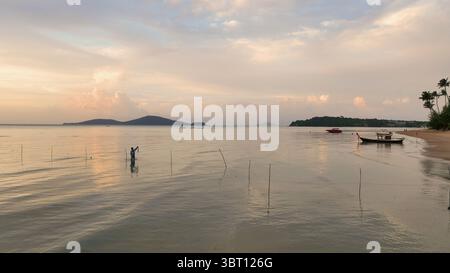 Vue aérienne d'un pêcheur solitaire jette son filet dans les eaux tranquilles et chatoyantes reflétant le ciel pastel près de la rive, Phuket, Phuket, Thaïlande. Banque D'Images
