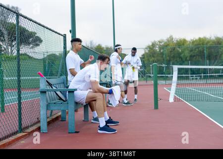 Divers coéquipiers masculins assis et debout sur un court de tennis tenant des raquettes, serviette et bouteille d'eau Banque D'Images