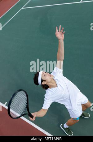 Joueur de tennis asiatique servant la balle sur un court dur extérieur avec raquette de tennis et bandeau blanc Banque D'Images