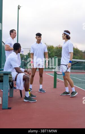 Divers coéquipiers masculins parlant tactiques tout en se reposant sur le banc avec serviette et bouteille près du filet de tennis Banque D'Images