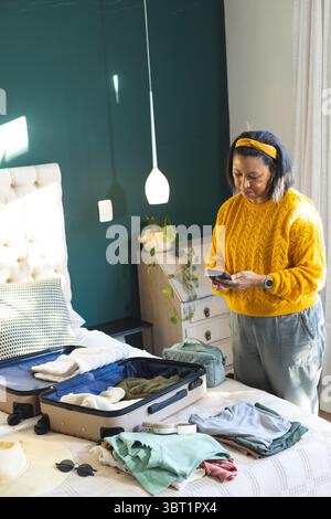 Femme afro-américaine mature organisant des vêtements dans une valise, vérifiant le téléphone dans la chambre à coucher, espace de copie Banque D'Images