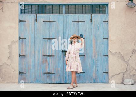 Jeune femme en robe florale et chapeau cliquant sur des photos au quartier historique Al seef à Dubaï, elle est entourée par l'architecture émiratie et arabe Banque D'Images