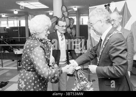 Présentation des croix de résistance Beverwijk présentation Beverwijk pays-Bas, Whizgle News, Dutch Desk, pays-Bas, 1950 - 2000 le 29-04-1983. L'image inclut ces rubriques. Dans un cadre intérieur animé, un groupe d'individus est engagé dans un moment d'interaction. Le fond comporte une bannière proéminente et plusieurs plantes en pot qui ajoutent une touche de verdure à la scène. Au premier plan, une femme aux cheveux bouclés et blancs, portant une robe à motifs aux tons sombres, sourit chaleureusement alors qu'elle tend sa main vers un homme habillé en tenue formelle, avec une ceinture et une chaîne de O. Banque D'Images