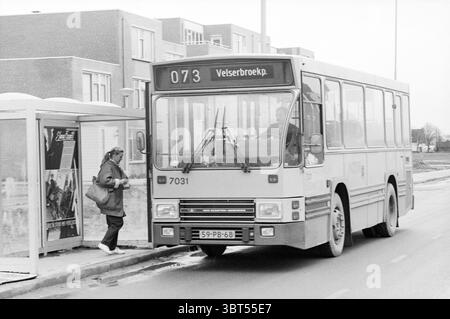 Bus Velserbroek P. [Velserbroekpolder] bus bus tours Velserbroek, Whizgle News, Dutch Desk, pays-Bas, 1950 - 2000 on 27-11-1987. L'image contient ces rubriques. La scène capture un moment à un arrêt de bus où un bus urbain est positionné, sa face avant faisant légèrement face au spectateur. Le bus est principalement d'une couleur pâle, se démarquant sur un ciel gris et couvert. Son design reflète une esthétique utilitaire, avec des lignes épurées et une forme carrée caractéristique des véhicules de transport public. À gauche du bus, une femme âgée descend du trottoir, vêtue d'un long manteau et d'une tête Banque D'Images