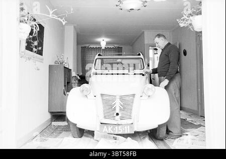 Man with his Ugly Duck, Whizgle News, Dutch Desk, pays-Bas, 1950 - 2000 en 1964. Ce sont les éléments de l'image. Dans une entrée étroite, une voiture blanche vintage occupe une position centrale, ses lignes courbes et son design rétro suggérant une esthétique du milieu du XXe siècle. Les murs du couloir sont ornés de lambris de bois de couleur claire, créant une atmosphère chaleureuse et accueillante. Un éclairage doux et diffus émane des plafonniers, créant une lueur douillette qui contraste légèrement avec l'extérieur lumineux de la voiture. Devant le véhicule, un homme d'âge moyen, vêtu d'un pull sombre, se penche légèrement f Banque D'Images