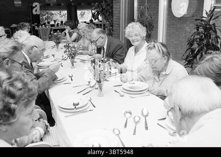 Table à manger ouverte Bennebroek nourriture et restaurants Bennebroek, Whizgle News, Dutch Desk, pays-Bas, 1950 - 2000 le 22-07-1988. L'image montre ces rubriques. Dans une salle à manger spacieuse baignée de lumière naturelle, une longue table s'étend sur la scène, ornée d'une nappe blanche. La table est dressée pour un repas commun, chaque cadre présentant des assiettes blanches, des couverts en argent étincelant et de la verrerie. Autour de la table, un groupe diversifié de personnes âgées est réuni, leurs expressions reflétant un mélange d’engagement et de nostalgie. Certains se penchent sérieusement vers l'avant, tandis que d'autres se concentrent intensément sur le Banque D'Images