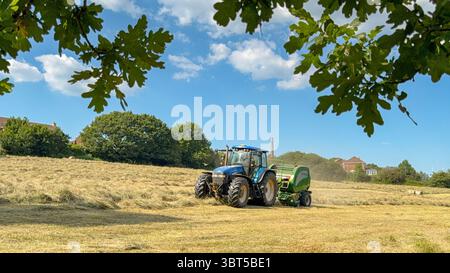Pontypridd, pays de Galles, Royaume-Uni - 12 juillet 2025 : tracteur remorquant une machine de pressage pour fabriquer de grandes balles rondes à partir d'herbe séchée dans un champ agricole. Banque D'Images
