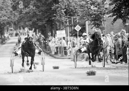 Course sur piste courte Zandvoort Zandvoort, Whizgle News, Dutch Desk, pays-Bas, 1950 - 2000 le 18-07-1991. Ces rubriques sont illustrées dans l'image. La scène capture une course de trot animée dans une rue bordée de spectateurs. Deux chevaux, tirant chacun un sulky, sont en plein mouvement, des sabots poussant un léger nuage de poussière de la piste de terre. Sur la gauche, un cheval à revêtement foncé avec un harnais audacieux et vibrant arbore un couvre-chef rouge et blanc saisissant conçu pour ressembler au visage d'un animal ludique, ajoutant une touche fantaisiste à la compétition. Le conducteur, habillé dans un costume confortable orné de motifs lumineux, m Banque D'Images