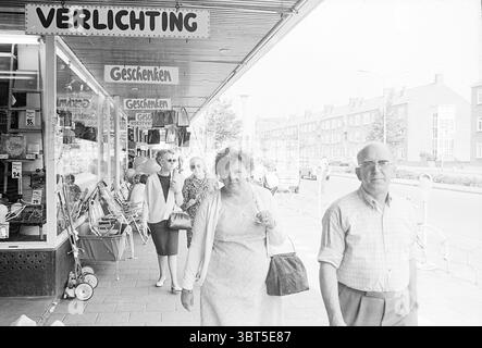 Shopping public IJmuiden Lange Nieuwstraat pays-Bas, Whizgle News, Dutch Desk, pays-Bas, 1950 - 2000 sur 06-1970. L'image montre ces rubriques. La scène capture un trottoir urbain animé, caractérisé par une zone commerçante remplie d'une variété de magasins. Le premier plan montre un couple marchant étroitement ensemble ; la femme, aux cheveux bouclés volumineux, porte une robe de couleur claire qui tombe juste en dessous du genou, lui donnant une apparence classique. Elle porte un sac à main, suggérant qu'elle aurait pu faire des courses. À côté d'elle, l'homme porte des lunettes et est habillé d'une chemise à carreaux, emb Banque D'Images