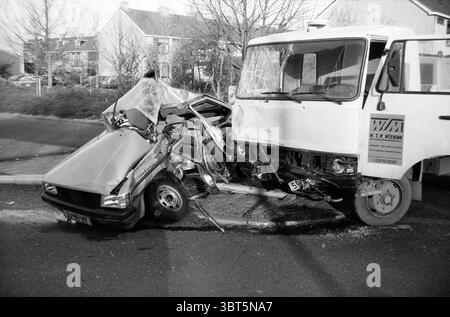 Collision d'une voiture de tourisme et d'un véhicule utilitaire de la société de transformation du béton W.T.M. Weenink, Whizgle News, Dutch Desk, pays-Bas, 1950 - 2000 sur 04-1985. L'image contient ces rubriques. La scène capture un moment de séquelles et de chaos. Au premier plan, une voiture compacte est sévèrement froissée, son capot plié vers le haut et son extrémité avant s'est écrasée sur le côté d'un grand camion de livraison blanc. Les deux véhicules sont garés sur une route bordée d'arbres clairsemés et sans feuilles, ajoutant une silhouette frappante sur un ciel couvert qui suggère une atmosphère sombre. La voiture, peinte dans un ton sombre, montre des signes de sh Banque D'Images