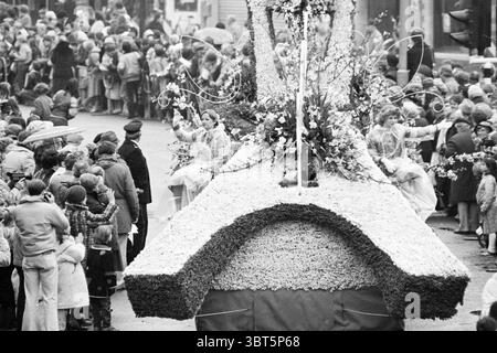 Défilé de fleurs Haarlem fleurs et filles de fleurs, Whizgle News, Dutch Desk, pays-Bas, 1950 - 2000 le 26-04-1980. Ces rubriques sont illustrées dans l'image. La scène capture un défilé animé en plein essor, caractérisé par une ambiance vibrante et festive, malgré les tons monochromes. À l'avant-garde, un grand flotteur floral se distingue, orné d'un arrangement complexe de fleurs et de verdure, suggérant des thèmes de célébration ou de printemps. Le design du flotteur présente des courbes douces et est décoré somptueusement, transmettant un sens artistique. Sur le char, deux individus vêtus de costumes élégants Banque D'Images