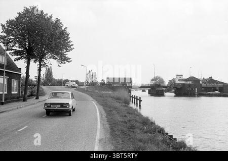 Pont sur le Ringvaart Oude Wetering (lac d'antan) ponts construction de ponts Oude Wetering, Whizgle News, Dutch Desk, pays-Bas, 1950 - 2000 sur 19-05-1987. Voici les rubriques de l'image. Une route sinueuse courbe doucement le long du bord de l’eau, bordée par un canal tranquille. D'un côté, une voiture classique se déplace sur la route, son design vintage évoquant une époque antérieure. La voiture est de couleur claire, contrastant avec les tons discrets de l'environnement. À gauche, un modeste bâtiment présente une façade simple avec quelques fenêtres, peut-être une résidence ou un petit magasin, tandis qu'un arbre avec lus Banque D'Images