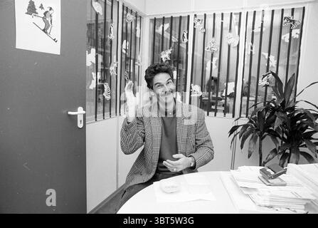 Martin Kooij Rijsenhout Rijsenhout, Whizgle News, Dutch Desk, pays-Bas, 1950 - 2000 le 11-02-1991. Ces rubriques sont illustrées dans l'image. La scène représente un homme assis à une table ronde, apparaissant animé alors qu'il fait des gestes avec une main levée. Il a une moustache distinctive et un comportement décontracté, portant un blazer sur une chemise rouge, ce qui ajoute une touche de personnalité à sa tenue. Autour de lui se trouvent divers éléments qui créent un environnement de bureau décontracté. Les murs autour de lui sont ornés de décorations ludiques, avec des figures découpées qui donnent une ambiance fantaisiste au décor. Une potte Banque D'Images