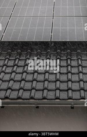 Panneaux solaires sur le toit en tuiles sombres pendant les fortes pluies avec gouttière et mur visibles. Banque D'Images