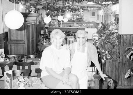 Restaurant italien IJmuiden IJmuiden pays-Bas, Whizgle News, Dutch Desk, pays-Bas, 1950 - 2000 le 19-05-1992. Ces rubriques apparaissent dans l'image. La scène capture un intérieur de restaurant confortable, caractérisé par une ambiance accueillante. Une femme est assise sur le bord d'une table en bois, vêtue d'une chemise blanche ample et d'un short, ses cheveux courts et bouclés contrastant avec sa tenue lumineuse. Elle a une expression joyeuse, rayonnant de chaleur et de convivialité. À côté d'elle se tient un homme portant un tablier de couleur claire au-dessus d'une chemise à col, présentant une position accueillante. Ses cheveux légèrement grisonnés et Banque D'Images