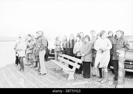 Arrivée d'un couple avec voilier de Cape Town Ships, Whizgle News, Dutch Desk, The Netherlands, 1950 - 2000 le 15-11-1974. Ce sont les éléments de l'image. La scène capture un rassemblement de personnes le long d'un front de mer. Les individus sont disposés en une ligne légèrement décalée, certains debout et d'autres assis sur un banc en bois. Il y a un mélange de styles de vêtements, typiques d'un climat plus frais, avec de nombreux manteaux, foulards et chapeaux portant, suggérant une atmosphère vigoureuse. Le cadre comporte une surface pavée, probablement faite de briques imbriquées, créant un sol texturé qui mène au w Banque D'Images