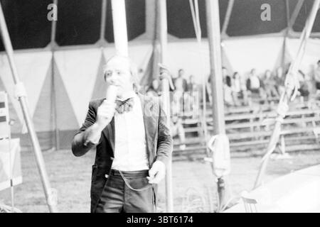 Cours de reportage Kimmie H'dorp cours de reportage, Whizgle News, Dutch Desk, pays-Bas, 1950 - 2000 le 09-06-1977. L'image inclut ces rubriques. La scène capture une atmosphère de cirque animée sous une grande tente. Une figure centrale, un homme vêtu d'un costume noir classique avec un noeud papillon, se tient bien en vue au premier plan, engageant de manière animée le public. Il tient un micro, suggérant qu'il présente une performance ou interagit avec les spectateurs. La toile de fond est remplie d'un mélange de motifs hexagonaux créés par la structure de la tente, éclairés par une lumière chaude et diffuse Banque D'Images