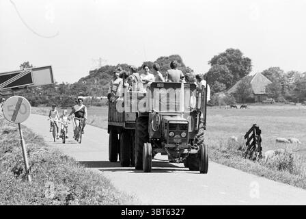 Start Polderloop Spaarnwoude Run Start Spaarnwoude, Whizgle News, Dutch Desk, pays-Bas, 1950 - 2000 le 07-07-1985. L'image inclut ces rubriques. La scène se déroule sur une route rurale qui s'étend à travers une campagne verdoyante. Une journée lumineuse et ensoleillée projette une lueur chaude sur le paysage, améliorant la vivacité des couleurs. Au premier plan, un tracteur bleu robuste tire une remorque en bois remplie de balles de foin. L'apparence altérée du tracteur suggère qu'il a connu de nombreuses saisons de travail acharné. Assis sur la remorque sont plusieurs personnes appréciant la balade, certains riant et bavardant, tandis que OTH Banque D'Images
