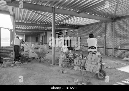Construction hockey sur glace - centre de tennis de table Zoetermeer chantiers de construction patinoire hockey sur glace Zoetermeer, Whizgle News, Dutch Desk, pays-Bas, 1950 - 2000 on 11-10-1982. Ces rubriques apparaissent dans l'image. La scène met en valeur un chantier de construction qui dégage un sens de l'industrialisation et du développement. Deux individus sont activement engagés dans leurs tâches, positionnés dans une structure partiellement construite avec des murs exposés en briques rectangulaires, suggérant une fondation robuste dans des tons chauds d'orange et de brun. À gauche, un ouvrier se penche vers le bas, se concentre sur la pose de briques, Banque D'Images