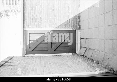 Subsidence du trottoir aux maisons Schalkwijk et Boerhaavewijk, Whizgle News, Dutch Desk, pays-Bas, 1950 - 2000 on 30-05-1967. Voici les rubriques de l'image. La scène présente une porte en bois rustique légèrement entrouverte, encadrée par un mur de briques rugueux et texturé. La porte a un ton profond et terreux, caractérisé par des motifs de grain visibles et un design simpliste avec des lattes diagonales. Le mur environnant affiche une teinte rougeâtre altérée, probablement de vieilles briques, créant un contraste chaleureux avec la porte plus sombre. Le sol est composé de pierres pavées, disposées selon un motif linéaire menant vers th Banque D'Images