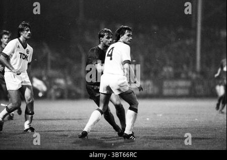 Football Haarlem - Ajax, Whizgle News, Dutch Desk, pays-Bas, 1950 - 2000 sur 09-1983. Ces rubriques apparaissent dans l'image. La scène capture un moment dans un match de football animé sous la lueur tamisée des lumières du stade. L'atmosphère est chargée d'énergie et d'excitation. Au premier plan, deux joueurs sont engagés dans un échange compétitif. Un joueur, vêtu d'un maillot sombre, semble affirmé alors qu'il s'approche d'un autre en uniforme blanc, qui pivote vers l'extérieur. Le joueur en blanc, avec le chiffre '4' affiché bien en évidence sur son dos, est dans une position dynamique, suggérant qu'il se prépare à déplacer Q. Banque D'Images