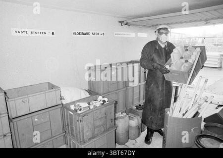 Responsable du dépôt de déchets chimiques d'entrepôt Hillegom Chemistry Hillegom, Whizgle News, Dutch Desk, pays-Bas, 1950 - 2000 on 05-12-1989. Ces rubriques sont illustrées dans l'image. La scène représente une zone de stockage encombrée, probablement un atelier ou un espace industriel. Diverses caisses et conteneurs robustes et altérés par les intempéries sont empilés et dispersés, certains remplis d'un assortiment d'articles. Les couleurs sont atténuées, avec une prédominance de gris et de tons terreux, créant une atmosphère industrielle et utilitaire. Une personne se tient bien en vue au premier plan, portant un équipement de protection qui comprend un masque et des lunettes Banque D'Images