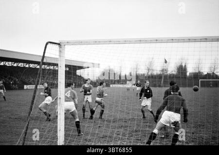 Football match., Whizgle News, Dutch Desk, pays-Bas, 1950 - 2000. Ces rubriques apparaissent dans l'image. La scène capture un moment dynamique dans un match de football, avec pour toile de fond un ciel nuageux et couvert qui projette une lumière silencieuse sur le terrain. L'herbe semble légèrement usée et boueuse, indiquant un jeu bien joué. Au premier plan, le cadre de but se tient bien en évidence, son filet oscillant légèrement comme s'il était perturbé par une action récente. Un joueur au premier plan, avec un maillot et un short, est concentré sur le ballon qui approche, accroupi et prêt à l'action. Il porte un uniforme sombre, Banque D'Images