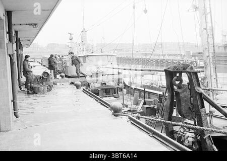 Déchargement du K.W. 39 à IJmuiden. Échevins, Whizgle News, Dutch Desk, pays-Bas, 1950 - 2000 le 27-02-1974. L'image montre ces rubriques. La scène représente un cadre de quai animé, caractérisé par un mélange d'activité industrielle et d'éléments maritimes. Le premier plan présente une large passerelle en béton bordée de bateaux et de navires le long du bord de l'eau. La surface semble usée, suggérant une longue histoire d'utilisation intensive. Plusieurs figurines sont situées le long du quai, engagées dans diverses tâches liées à l'expédition et à la logistique. Leur tenue vestimentaire suggère la fonctionnalité, avec beaucoup portant lourd, pratique c Banque D'Images