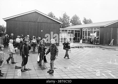 Terrain de jeu Mearten van Heemskerckschool. Terrains de jeux et écoles de terrains de jeux, Whizgle News, Dutch Desk, pays-Bas, 1950 - 2000 on 09-09-1974. L'image montre ces rubriques. Dans une scène représentant une cour d'école, un groupe diversifié d'enfants et d'adultes est réuni, créant une atmosphère animée. Le décor présente deux structures en bois au design simple et utilitaire, caractérisées par un revêtement brun foncé. Ces bâtiments sont fonctionnels, avec de nombreuses fenêtres qui reflètent un ciel légèrement couvert. Le sol est fait de grandes tuiles carrées, certaines scintillantes de la pluie récente, améliorant le s. Banque D'Images
