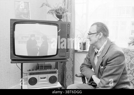 Émission de télévision russe Haarlem Television Haarlem pays-Bas, Whizgle News, Dutch Desk, pays-Bas, 1950 - 2000 le 13-03-1984. L'image contient ces rubriques. Dans un salon confortable et modeste, un homme plus âgé est assis sur un fauteuil à motifs, son regard fixé intentionnellement sur une télévision vintage. Il a une expression réfléchie, soulignée par l'éclairage doux et diffus qui filtre à travers une fenêtre voisine. La salle porte une ambiance nostalgique, avec des murs ornés d'un papier peint texturé et lumineux. La télévision, qui rappelle une époque révolue, affiche une scène floue en noir et blanc avec deux Banque D'Images