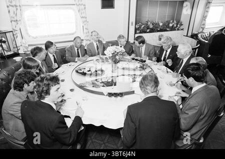 Hitachi Venture (poutre porteuse) IJmuiden Hoogovens IJmuiden pays-Bas, Whizgle News, Dutch Desk, pays-Bas, 1950 - 2000 on 16-04-1982. L'image inclut ces rubriques. Dans un espace salle à manger bien éclairé et élégamment aménagé, une grande table circulaire est remplie d'un assortiment de plats et de boissons. La nappe est d'un blanc immaculé, contrastant avec les teintes plus sombres du décor environnant. Une pièce maîtresse de fleurs fraîches ajoute une touche de couleur, tandis que des assiettes de divers plats, y compris probablement des salades et des pâtisseries, sont stratégiquement placées autour de la table. Assis autour de la table se trouvent un groupe d'hommes Banque D'Images