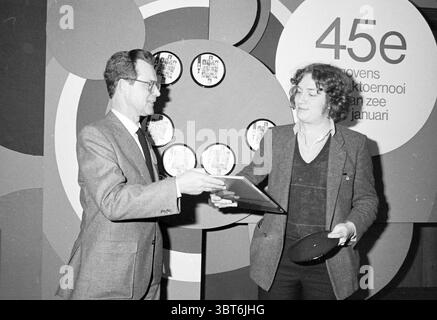 Joueurs d'échecs + Side Events Blast Furnace Chess Tournament, Whizgle News, Dutch Desk, pays-Bas, 1950 - 2000 le 13-01-1983. L'image contient ces rubriques. La scène capture un moment d’échange entre deux individus. A gauche, un homme vêtu d'une tenue habillée, avec des lunettes, remet un objet à l'homme à droite, qui a une apparence plus décontractée, vêtu d'un pull sombre sur une chemise boutonnée. Le destinataire semble engagé et légèrement joyeux, avec un soupçon de sourire, suggérant une interaction positive. En arrière-plan, un design vibrant dispose d'un grand Mo circulaire Banque D'Images