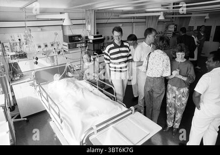 Open day intensive Care EC, Whizgle News, Dutch Desk, pays-Bas, 1950 - 2000 on 11-09-1990. Voici les rubriques de l'image. La scène se déroule dans une chambre d'hôpital, caractérisée par une disposition simple mais fonctionnelle. Au centre, un lit d'hôpital est entouré de matériel médical, avec un patient reposant sous une couverture de couleur claire. Le lit a des garde-corps de chaque côté, et divers moniteurs sont positionnés à proximité, affichant les signes vitaux. Autour du lit, un groupe de personnes se réunit, suggérant un moment de soutien et d’interaction. Certains se tiennent près, se penchant légèrement vers le lit, tandis que Banque D'Images