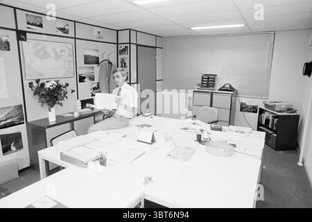 Fermeture de Lire Engineering dans Beyingsflat Persons Companies, Whizgle News, Dutch Desk, pays-Bas, 1950 - 2000 le 29-08-1989. Ces rubriques apparaissent dans l'image. La scène met en valeur un environnement de bureau caractérisé par une atmosphère professionnelle mais légèrement encombrée. La chambre présente une palette de couleurs principalement neutres, avec des murs blancs et une moquette grise. À gauche, un bureau est encombré de feuilles de papier, d'une pile de dossiers et de quelques objets personnels. Un arrangement floral ajoute une touche de couleur, avec des fleurs vibrantes qui se démarquent sur la toile de fond tamisée. Le bureau est accompagni Banque D'Images