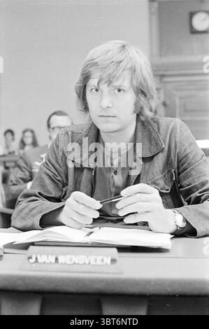 Portrait J. van Wensveen, Whizgle News, Dutch Desk, pays-Bas, 1950 - 2000 sur 09-1971. L'image montre ces rubriques. La scène montre un jeune homme assis à une table officielle dans un environnement sombre et professionnel, probablement une salle d'audience ou une chambre législative. Ses cheveux longs et ondulés sont clairs, ce qui lui donne une apparence un peu décontractée qui contraste avec la gravité du décor. Il porte une veste sombre sur une chemise boutonnée, laissant entendre un mélange d'informalité et de sérieux dans sa tenue. Dans ses mains, il tient une paire de lunettes, positionnée juste devant lui sur la table. Son expression Banque D'Images