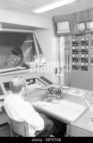 Salle de contrôle., Whizgle News, Dutch Desk, pays-Bas, 1950 - 2000. L'image montre ces rubriques. La scène représente une salle de contrôle vintage, caractérisée par son design du milieu du XXe siècle. Un individu solitaire est assis à un grand bureau incurvé, qui est équipé d'un ensemble de boutons, de cadrans et de leviers, suggérant un système complexe en fonctionnement. L'individu, vêtu d'une chemise formelle, fait face à une grande fenêtre en verre qui offre une vue de l'espace au-delà, bien que les détails de cet espace ne soient pas visibles. Le pupitre de commande a une surface en bois poli avec divers instruments stratégiques Banque D'Images