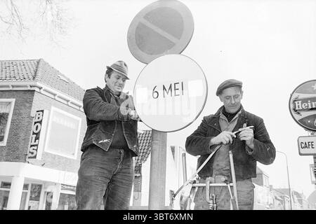 Mise en place de panneaux de signalisation trafic sécurité routière, Whizgle News, Dutch Desk, pays-Bas, 1950 - 2000 le 20-03-1969. L'image inclut ces rubriques. Dans cette scène, deux hommes sont engagés dans une tâche, debout devant un panneau circulaire qui est destiné à transmettre une date. Le signe est bien visible, attirant l'attention en raison de sa simplicité et de sa forme circulaire. Le premier homme, positionné à gauche, a une expression sérieuse car il se concentre intensément sur le signe, portant une veste sombre et une casquette qui ajoute à son apparence robuste. Il semble ajuster ou manipuler la rue du panneau Banque D'Images