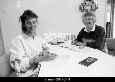MRS Trip from Amnesty International Hillegom Hillegom, Whizgle News, Dutch Desk, pays-Bas, 1950 - 2000 on 22-05-1990. L'image inclut ces rubriques. La scène capture deux individus assis à une table de couleur claire, qui sert de point focal central. La première personne, à gauche, a les cheveux foncés à la longueur des épaules et un doux sourire. Elle est habillée décontractée d'une veste blanche, qui contraste avec le fond sombre derrière elle. Ses mains sont placées sur une pile de papiers, indiquant son engagement dans une discussion ou une activité. Le deuxième individu, à droite, a des cheveux courts et gris bouclés Banque D'Images