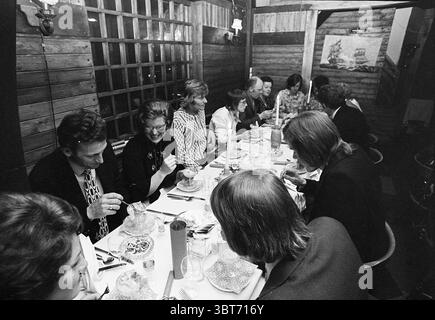 Célébration anniversaire etc, Whizgle News, Dutch Desk, pays-Bas, 1950 - 2000 le 04-1974. L'image inclut ces rubriques. La scène capture un rassemblement animé autour d'une longue table à manger, situé dans un espace chaleureux et rustique caractérisé par des boiseries et un éclairage tamisé. La table est ornée de vaisselle blanche aux motifs délicats, créant un contraste avec la surface en bois sombre. Engagé avec enthousiasme dans la conversation, un groupe diversifié de personnes occupe les deux côtés de la table. Ils sont habillés en tenue formelle, avec beaucoup de blazers et de robes qui contribuent un air de sophisme Banque D'Images