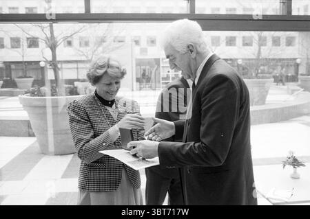 Médaille d'argent pour Mr. Niekerk Sports Council Presentation, Whizgle News, Dutch Desk, pays-Bas, 1950 - 2000 le 10-02-1990. L'image contient ces rubriques. Dans un cadre intérieur bien éclairé avec de grandes fenêtres qui permettent à la lumière naturelle d’inonder l’espace, il y a un moment d’interaction capté entre trois individus. L'arrière-plan présente un design architectural moderne avec des surfaces lisses en verre et en béton, encadrées par de la verdure extérieure dans de grandes jardinières qui fournissent un soupçon de vie. La figure centrale est une femme aux cheveux bouclés et au sourire éclatant, portant un blazer à motifs qui combine Sha Banque D'Images