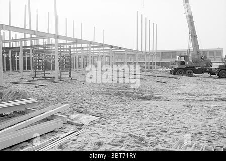 Construction de nouvelles maisons., Whizgle News, Dutch Desk, pays-Bas, 1950 - 2000. L'image contient ces rubriques. La scène présente un chantier caractérisé par une esthétique en noir et blanc qui met en valeur les textures et les formes des structures en construction. Au premier plan, une étendue sablonneuse est visible, parsemée de matériaux dispersés tels que des poutres métalliques et des planches de bois, indiquant les travaux en cours. Les colonnes verticales en acier s'élèvent en évidence vers le ciel, formant un squelette d'un bâtiment, suggérant les premières étapes de la construction. Les colonnes sont frappées contre le Banque D'Images