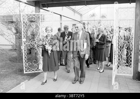 Ouverture Liliade Opening Openings Commissioning, Whizgle News, Dutch Desk, pays-Bas, 1950 - 2000 on 10-05-1979. L'image contient ces rubriques. La scène représente un moment de célébration alors qu'un groupe émerge à travers des portes en fer forgé blanc ornées. Les portes, décorées avec des motifs complexes, encadrent l'entrée d'un lieu qui a une sensation lumineuse et aérée en raison de la structure de verre environnante et de la verdure abondante. Au premier plan, un homme et une femme marchent côte à côte. La femme, vêtue d'un long manteau et tenant un bouquet de fleurs, dégage un air de joie et d'accomplissement. Son expression Banque D'Images