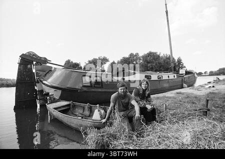 Couple pour personnes Tjalk, Whizgle News, Dutch Desk, pays-Bas, 1950 - 2000 on 11-09-1973. Ces rubriques apparaissent dans l'image. Dans cette scène tranquille au bord de l'eau, un couple s'assoit décontracté sur le bord d'un petit bateau en bois, leurs postures détendues suggérant une camaraderie confortable. L'homme, portant une chemise simple et un pantalon robuste, a un sourire facile alors qu'il se penche légèrement en avant. À côté de lui, une femme aux cheveux longs, vêtue d'une robe à motifs, semble engagée dans la conversation, ses yeux scintillant d'un sentiment de joie. Derrière eux, une barge plus grande et vintage repose doucement sur l'eau, sa peinture de coque Banque D'Images