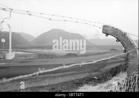 Montagnes de charbon de Hoogovens Hoogovens Velsen-Noord, Whizgle News, Dutch Desk, pays-Bas, 1950 - 2000 le 25-02-1982. L'image montre ces rubriques. La scène est dominée par un paysage industriel sombre, caractérisé par de grands monticules de sol sombre ou de gravier qui s'élèvent bien en évidence sur la toile de fond. Les tons sont atténués, principalement dans les nuances de gris, créant une atmosphère sombre et quelque peu désolée. Une clôture en fil de fer barbelé traverse le premier plan, son métal tranchant et torsadé contrastant fortement avec la terre du sol environnant. Cette barrière ajoute un sentiment de confinement ou de séparation Banque D'Images