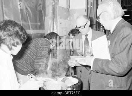Dégustation de vins chez Levie and Paarlberg Amsterdam Wine Amsterdam pays-Bas, Whizgle News, Dutch Desk, pays-Bas, 1950 - 2000 le 22-01-1980. Ces rubriques apparaissent dans l'image. Dans un cadre intérieur animé, un groupe d'individus s'engage autour d'une table chargée d'offres culinaires. La toile de fond présente un mélange de murs en bois non finis et de rideaux en plastique transparent, suggérant un environnement industriel ou temporaire où la préparation et l'échantillonnage des aliments ont lieu. Les gens sont divers en apparence ; une personne est penchée, apparemment inspectant ou préparant la nourriture, leur attention entièrement Banque D'Images